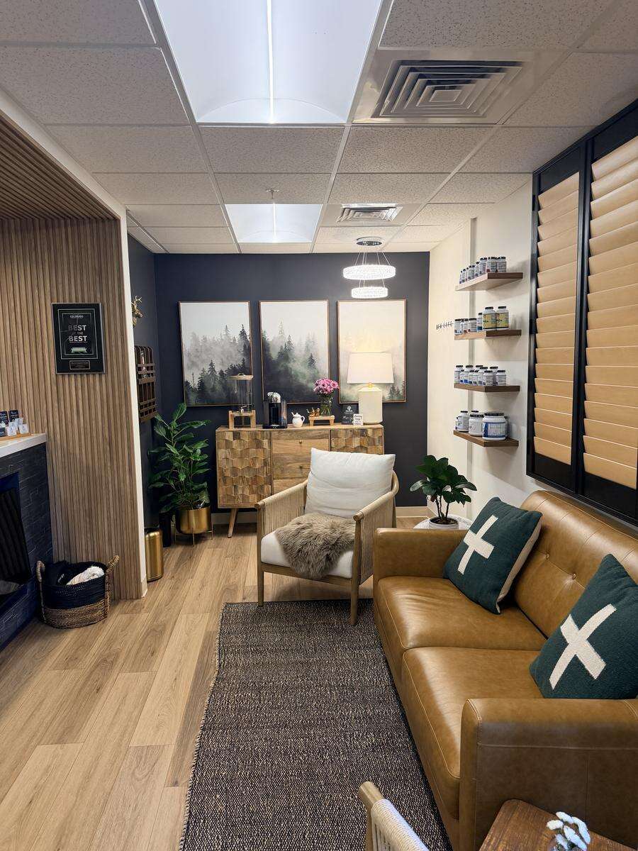 Integrative health clinic waiting room with tan leather sofa, woven accent chair, misty forest artwork on a navy accent wall, and wall shelves displaying supplements