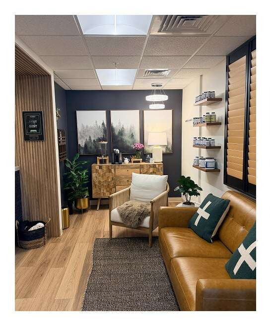 Integrative health clinic waiting room with tan leather sofa, woven accent chair, misty forest artwork, and wall shelves of supplements