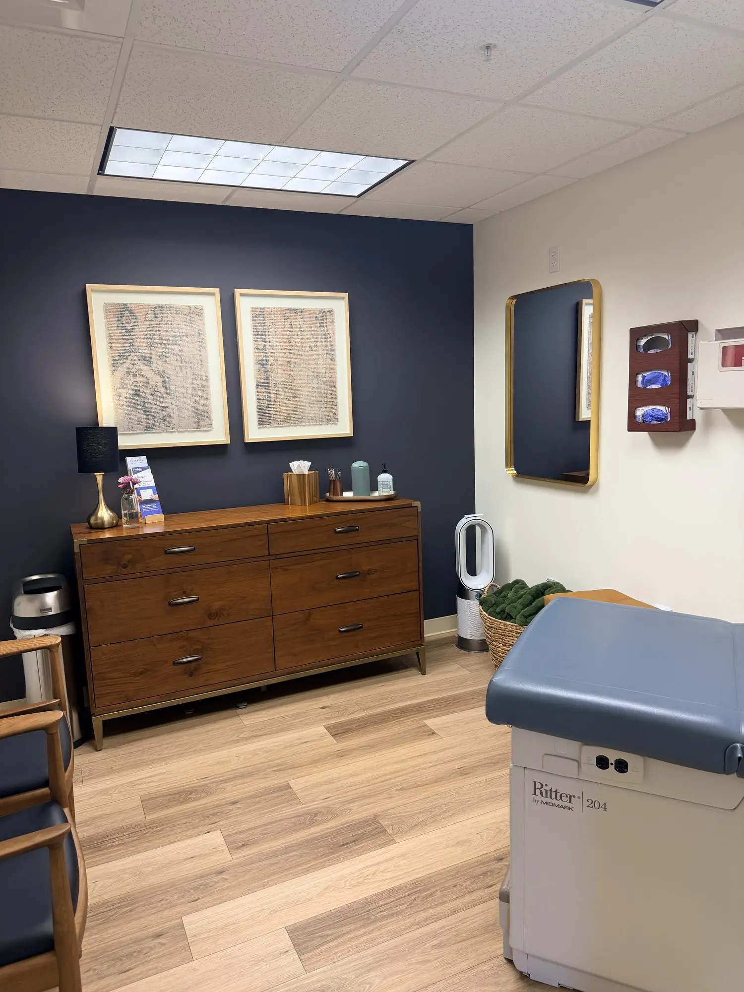 Medical exam room with navy accent wall, wood dresser, framed artwork, and a blue upholstered patient exam table