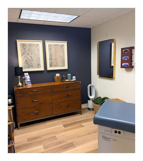 Medical exam room with navy accent wall, wood dresser, framed artwork, and a blue upholstered patient exam table