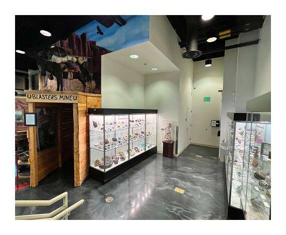 Museum gallery with illuminated gem and mineral display cases beside a rustic mine entrance exhibit