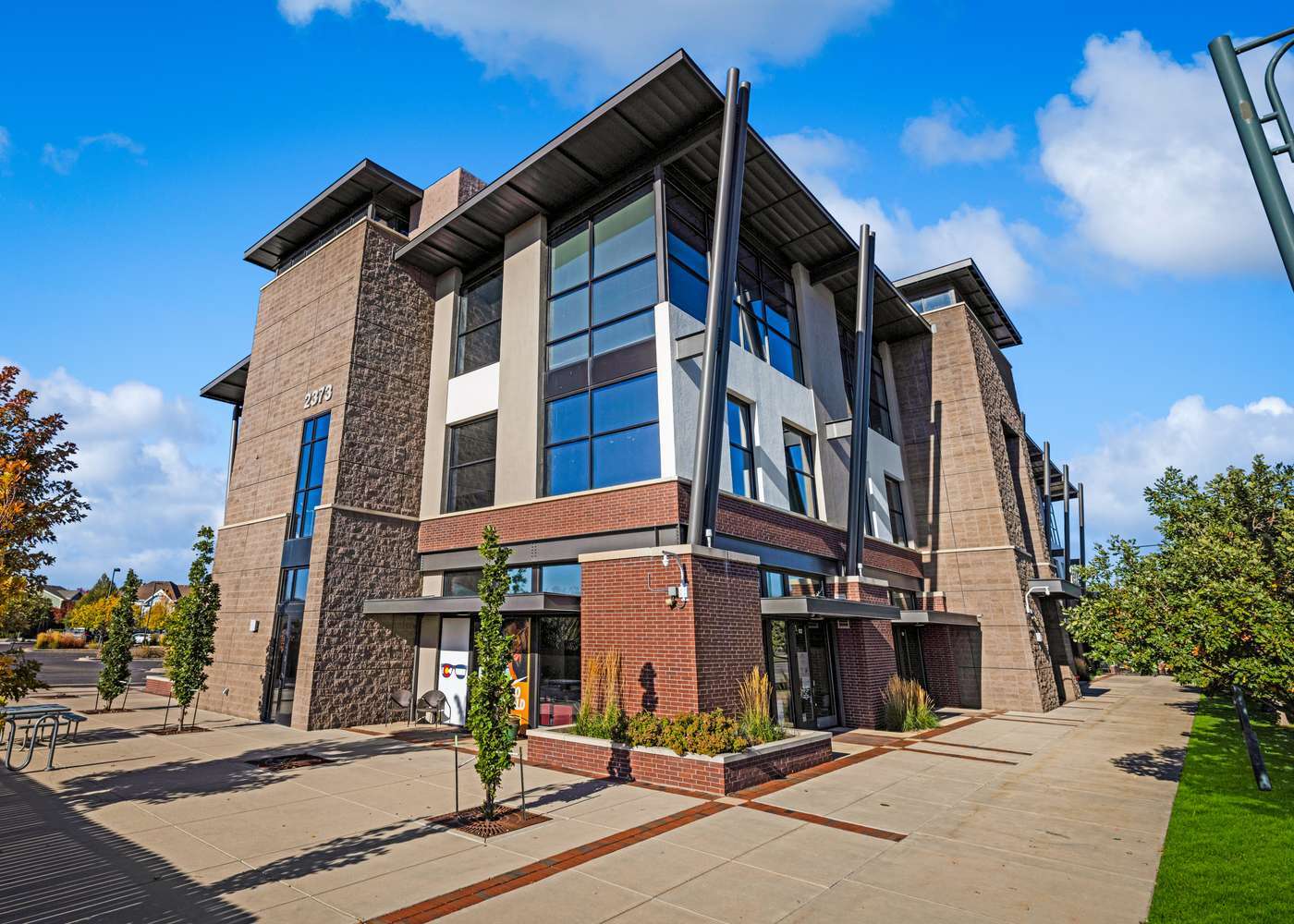 Three-story modern commercial office building with brick, stone, and stucco exterior, large tinted windows, and landscaped sidewalk entrance